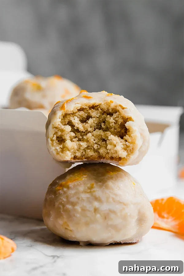 Two mandarin glazed donut holes, one with a bite taken out, showing the cakey interior, on a white plate with fresh mandarins in the background.