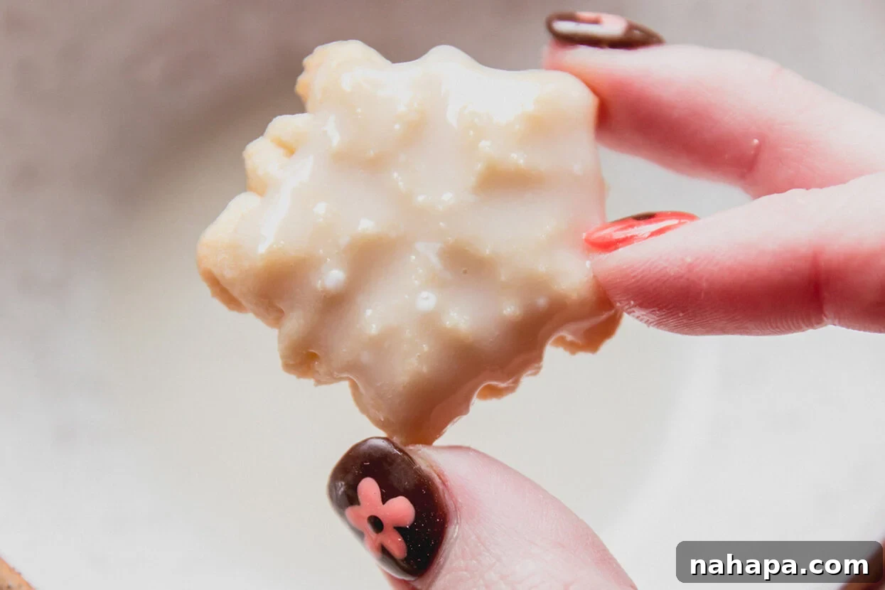 A delicate snowflake-shaped spritz cookie being dipped into a bowl of smooth white icing.