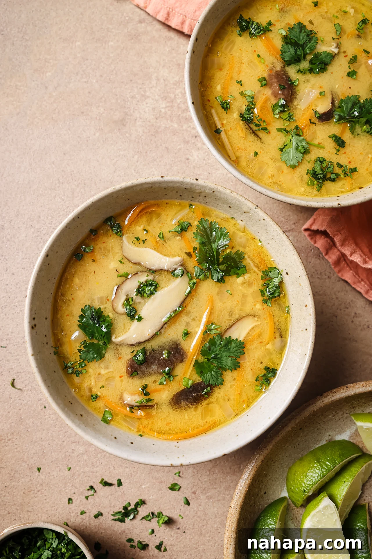 Two bowls of vibrant Tom Kha soup, garnished with fresh cilantro and lime wedges, ready to be served on a rustic wooden surface.