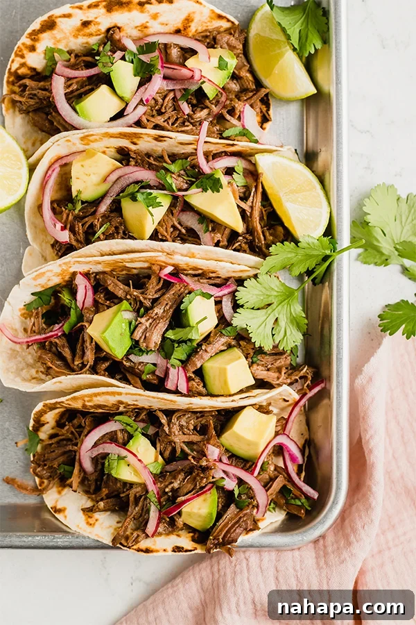 Shredded beef tacos topped with red onion, lime, and cilantro leaves on a white plate.