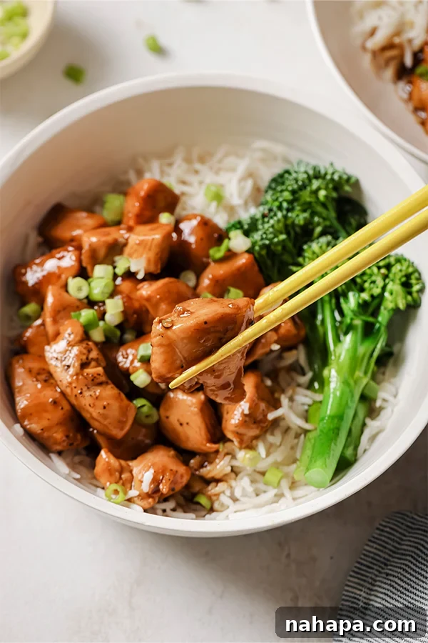 A close-up shot of a healthy bourbon chicken bowl, served with broccolini and rice noodles, with chopsticks poised to take a delicious bite, highlighting the texture and appeal of the meal.