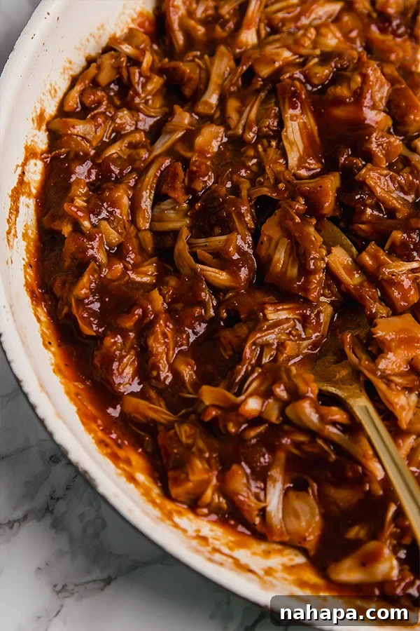 Smoky Pulled Jackfruit 3 A close-up shot of shredded BBQ jackfruit simmering in a rich, dark sauce in a pan, showcasing its texture and vibrant color.