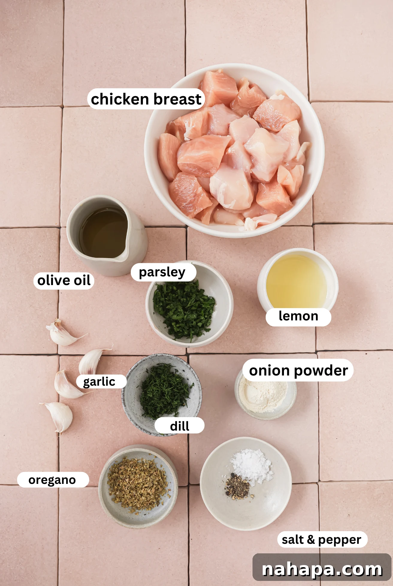 Fresh ingredients for chicken souvlaki marinade displayed in individual white bowls: olive oil, lemon, garlic, parsley, dill, onion powder, oregano, sea salt, and black pepper.