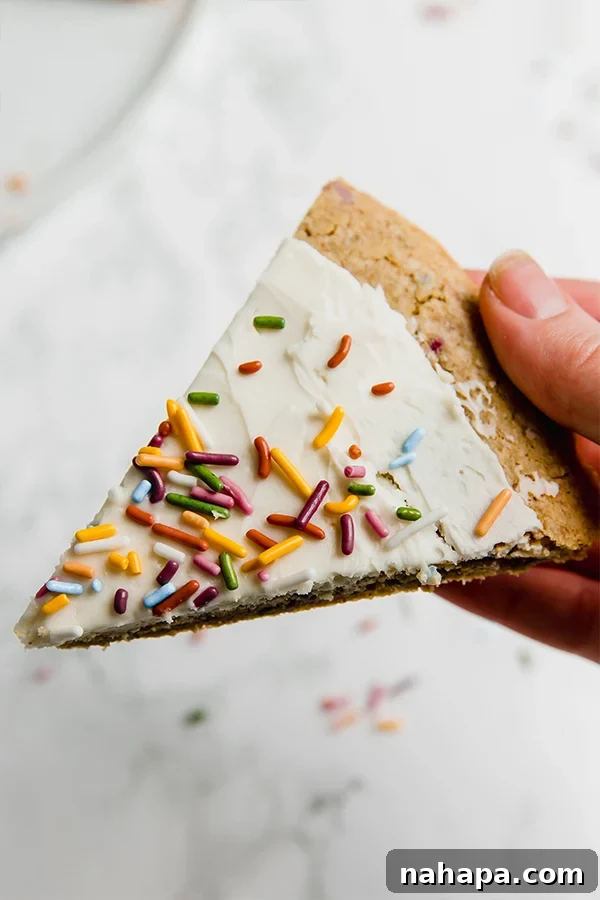 Close-up of a slice of funfetti cookie pizza, showing the thick frosting and colorful sprinkles, ready to be eaten.
