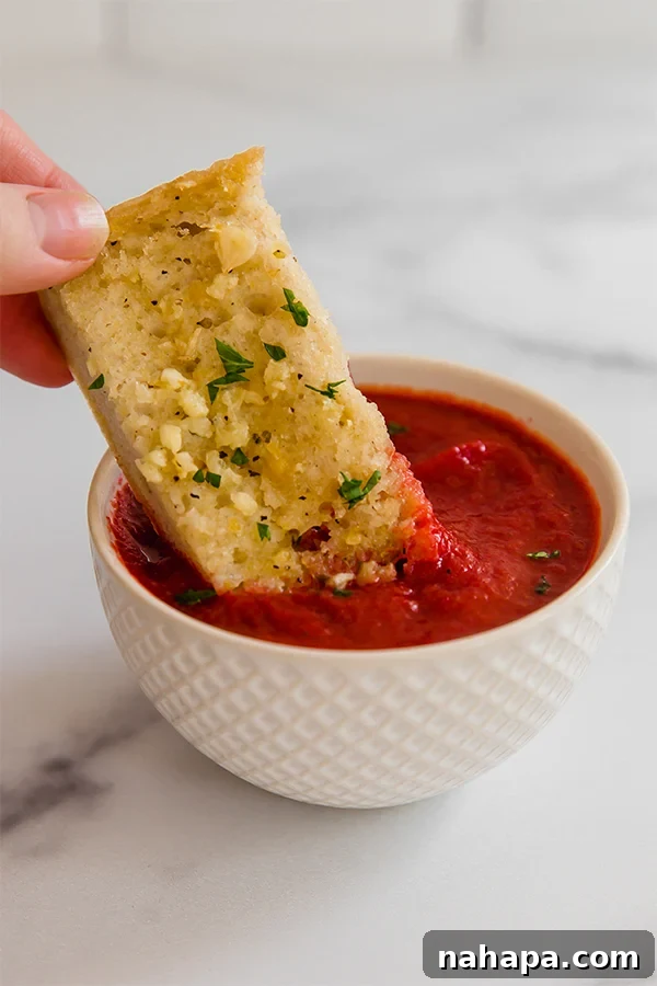 Close-up of a golden-brown gluten-free garlic bread slice being dipped into a small bowl of vibrant red marinara sauce, highlighting its crispy texture and delicious appeal.