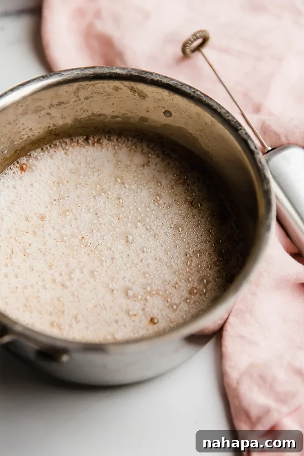 Step-by-step image showing almond milk, cinnamon, coconut sugar, and vanilla extract being combined in a small pot on a stovetop.