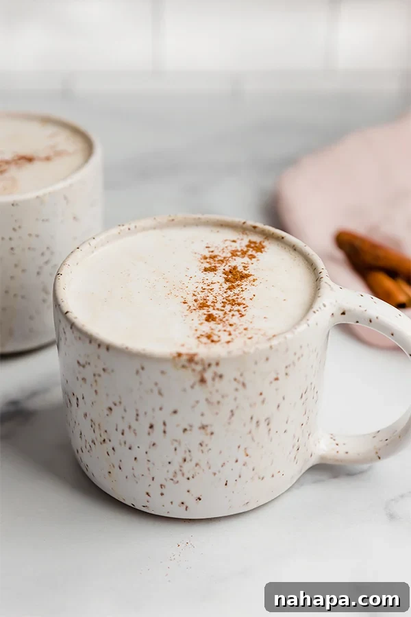 A top-down shot of a Snickerdoodle Latte in a clear mug, highlighting the creamy texture and cinnamon garnish, next to a small bowl of cinnamon sticks.