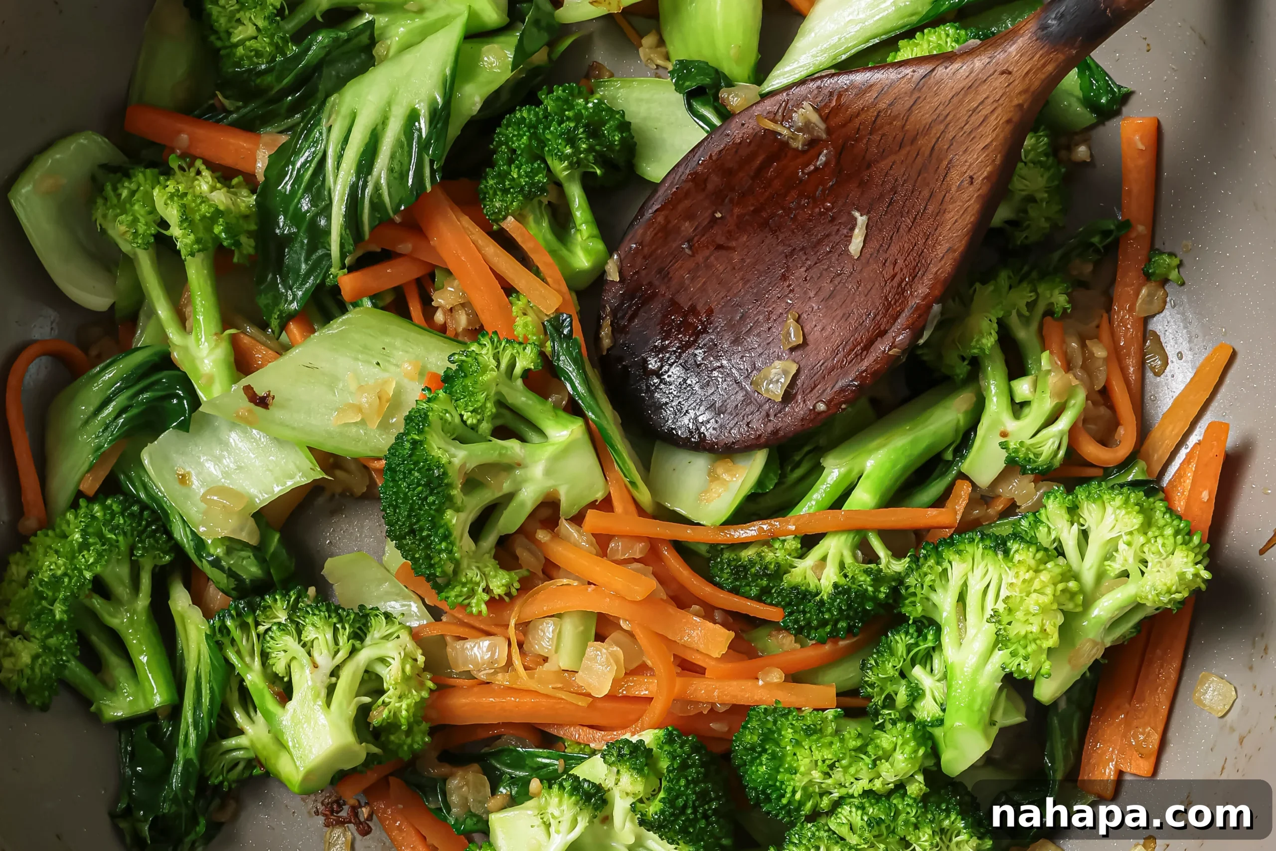 Sautéing diced onions and minced garlic in a hot pan with a wooden spoon, followed by adding a mix of broccoli, bok choy, and carrots.