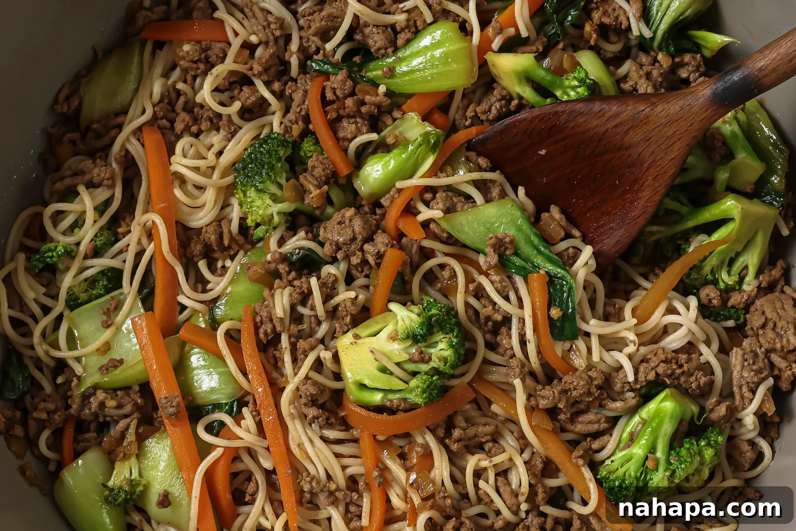 Combining cooked ground beef, sautéed vegetables, and softened ramen noodles in a large pan, mixing them with the stir-fry sauce.
