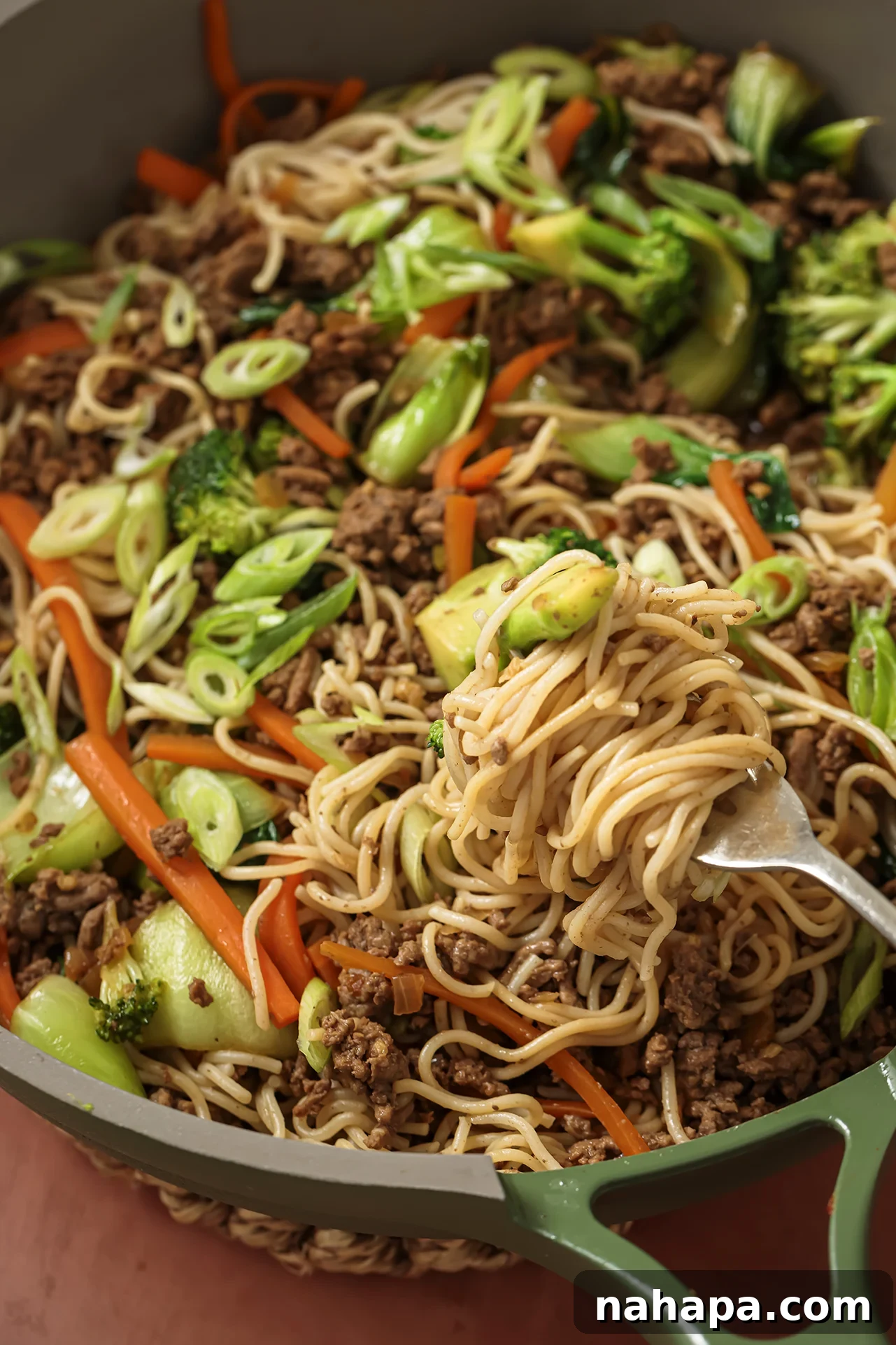 A close-up shot of a fork digging into a pan of ground beef ramen noodle stir fry, showcasing the rich texture, vibrant vegetables, and perfectly cooked noodles.