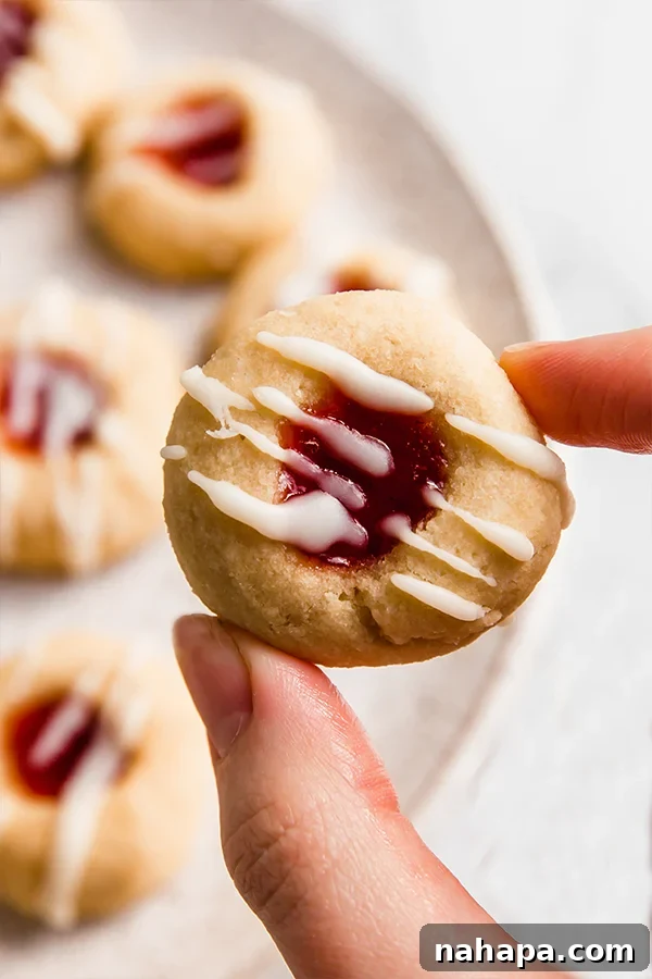 A close-up view of two thumbprint cookies on a plate, with one cookie delicately held between fingers, showcasing its jam-filled center and drizzle