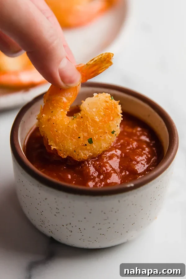 Oven-baked coconut shrimp being dipped into a bowl of cocktail sauce, showcasing its crispy texture