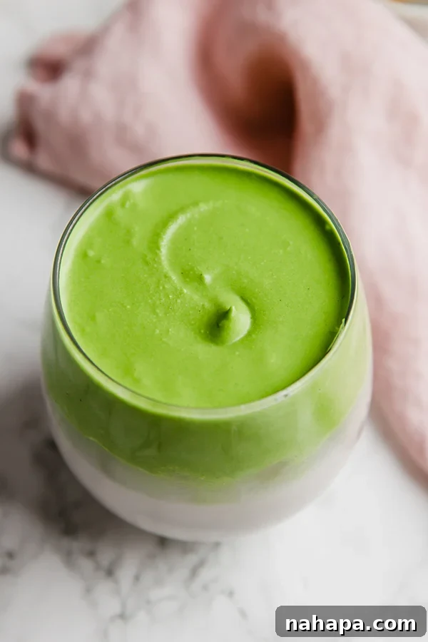 A vibrant green Dalgona Matcha drink served in a clear glass cup, showcasing its frothy whipped topping