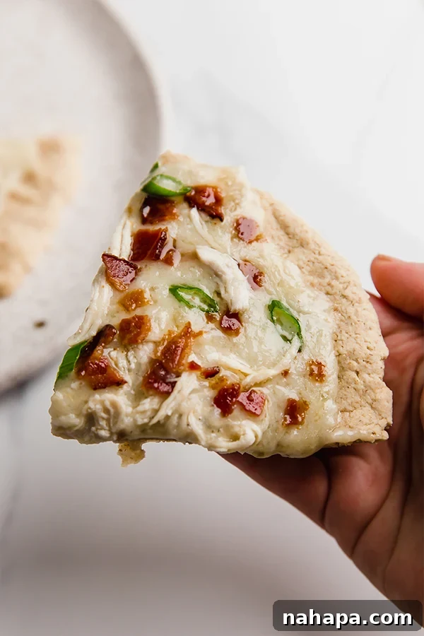 Hands holding a slice of AIP & Paleo Chicken Bacon Ranch Pizza, showing its satisfying texture and rich toppings
