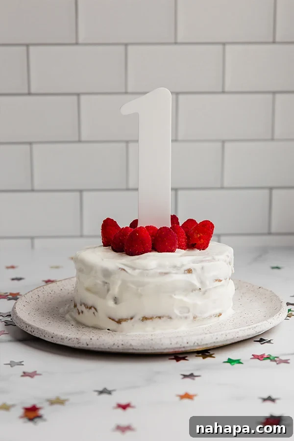 Joyful baby celebrating their first birthday with a colorful, healthy smash cake topped with fresh raspberries and a festive 'One' cake topper.