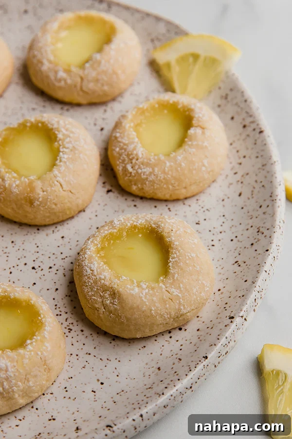 Close-up of baked lemon thumbprint cookies, lightly dusted with powdered sugar, on a white plate with fresh lemon slices.