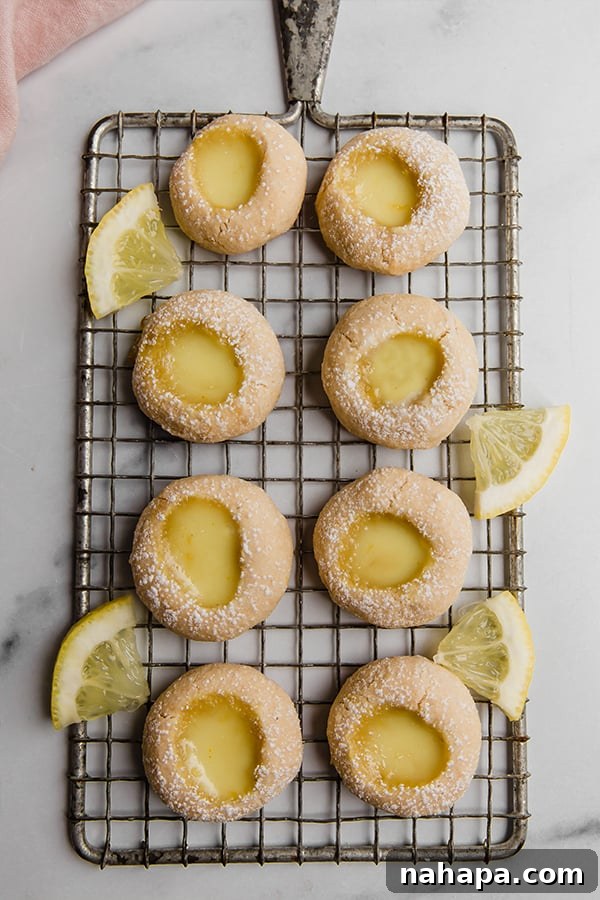 A stack of lemon bar thumbprint cookies on a plate, with a background of fresh lemons and green foliage, emphasizing their natural ingredients.