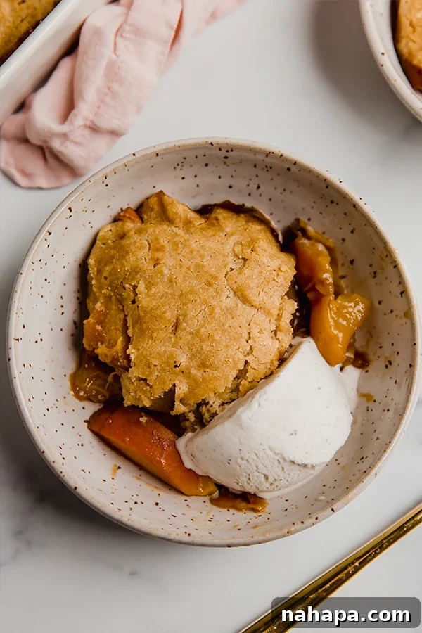 A top-down view of a steaming, cooked Paleo and AIP peach cobbler, glistening with a golden biscuit topping, served in a bowl with a generous scoop of vanilla dairy-free ice cream melting on its side.