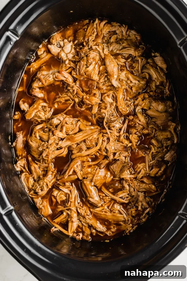 Tender BBQ pulled pork simmering in the insert of a slow cooker, surrounded by rich sauce, ready to be shredded.