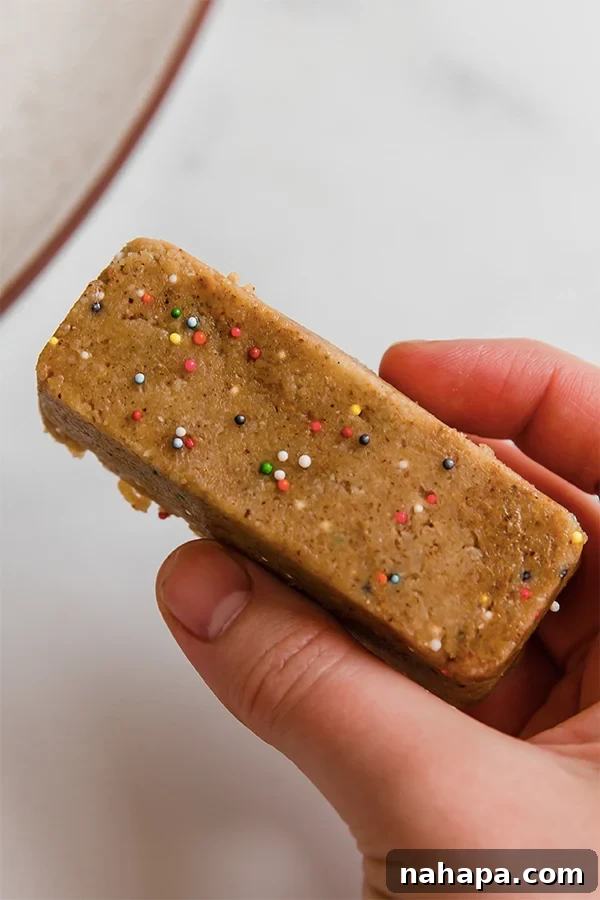 A single homemade cake batter protein bar, perfectly formed with colorful sprinkles, held gently in a hand, ready for a healthy and satisfying bite.