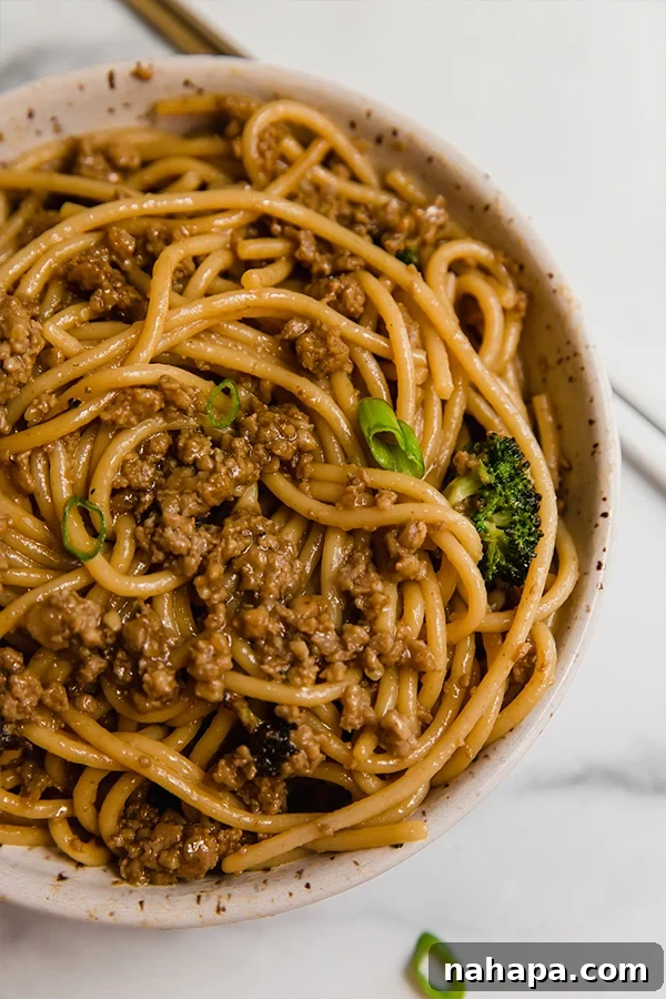 A bowl of Mongolian ground beef with noodles, garnished with green onions and fresh broccoli, served with chopsticks on a rustic wooden surface