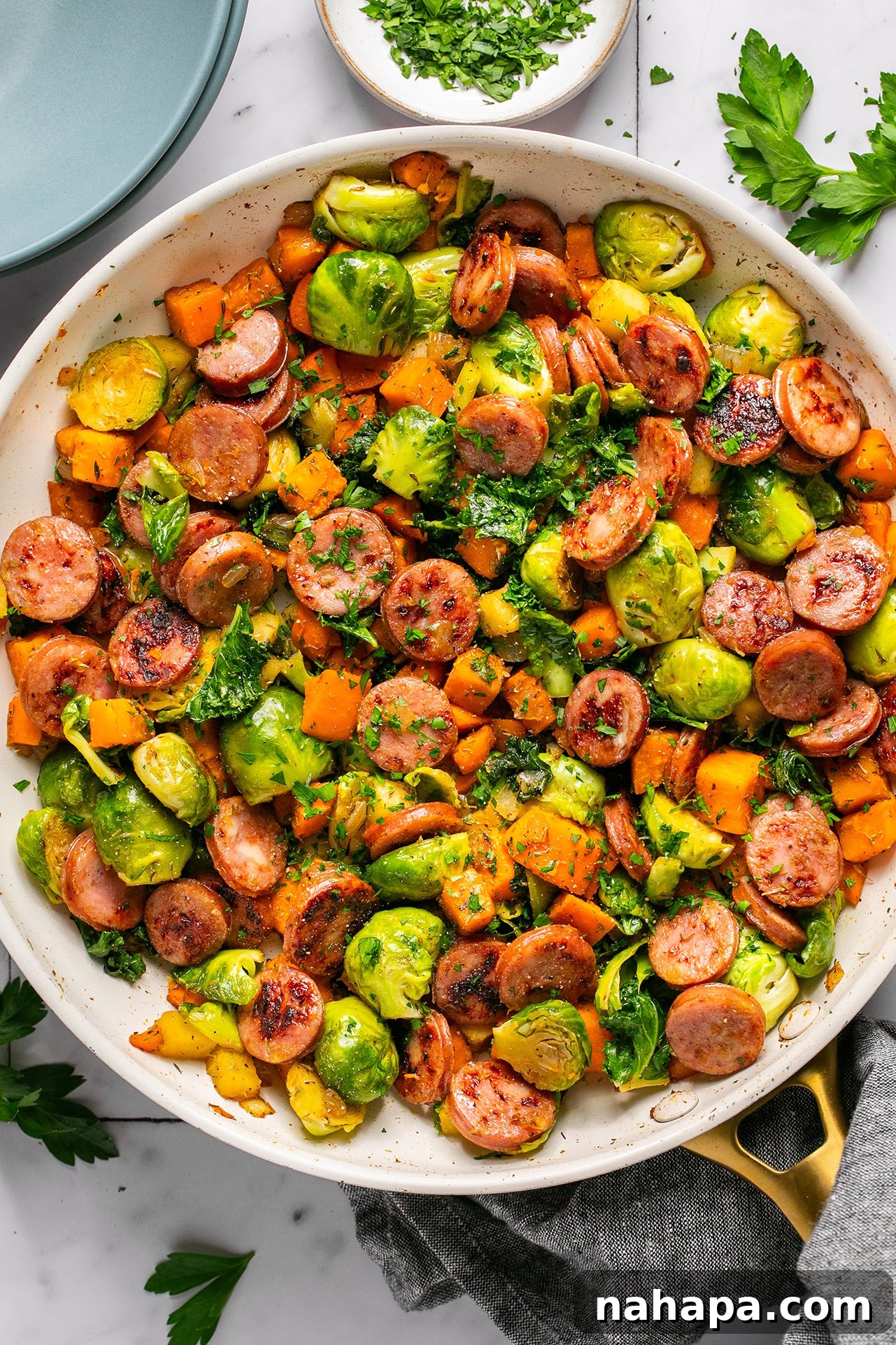 A large skillet filled with a colorful apple chicken sausage and vegetable mixture, including sliced sausage, diced sweet potatoes, halved Brussels sprouts, and apple pieces, garnished with fresh herbs.