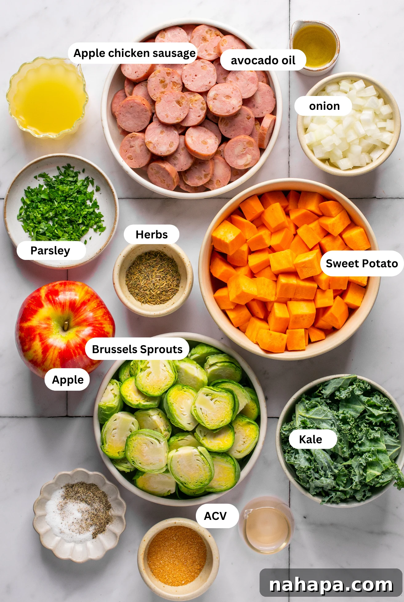 Various ingredients for an apple chicken sausage skillet laid out in separate bowls, including sliced sausage, diced sweet potato, halved Brussels sprouts, a diced Honeycrisp apple, fresh kale, and herbs.