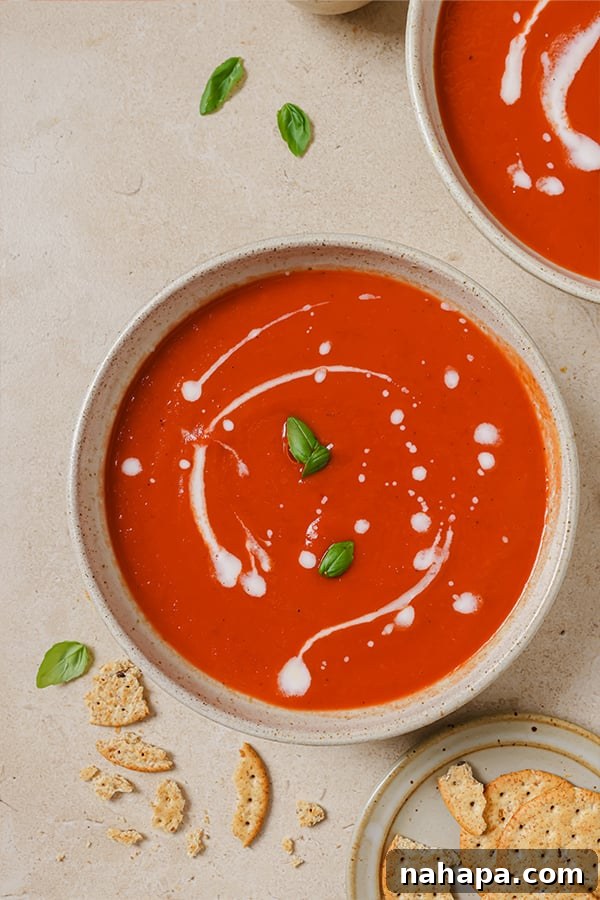 Two bowls of nightshade-free tomato soup on a wooden board with a spoon and herbs.