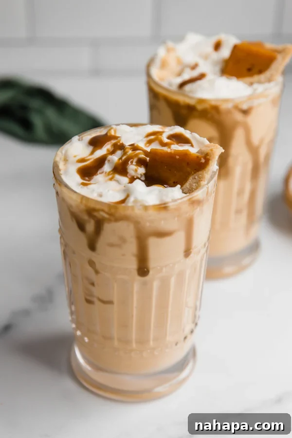 Two pumpkin pie milkshakes with generous whipped cream and pumpkin pie slices on top, against a rustic autumn background.