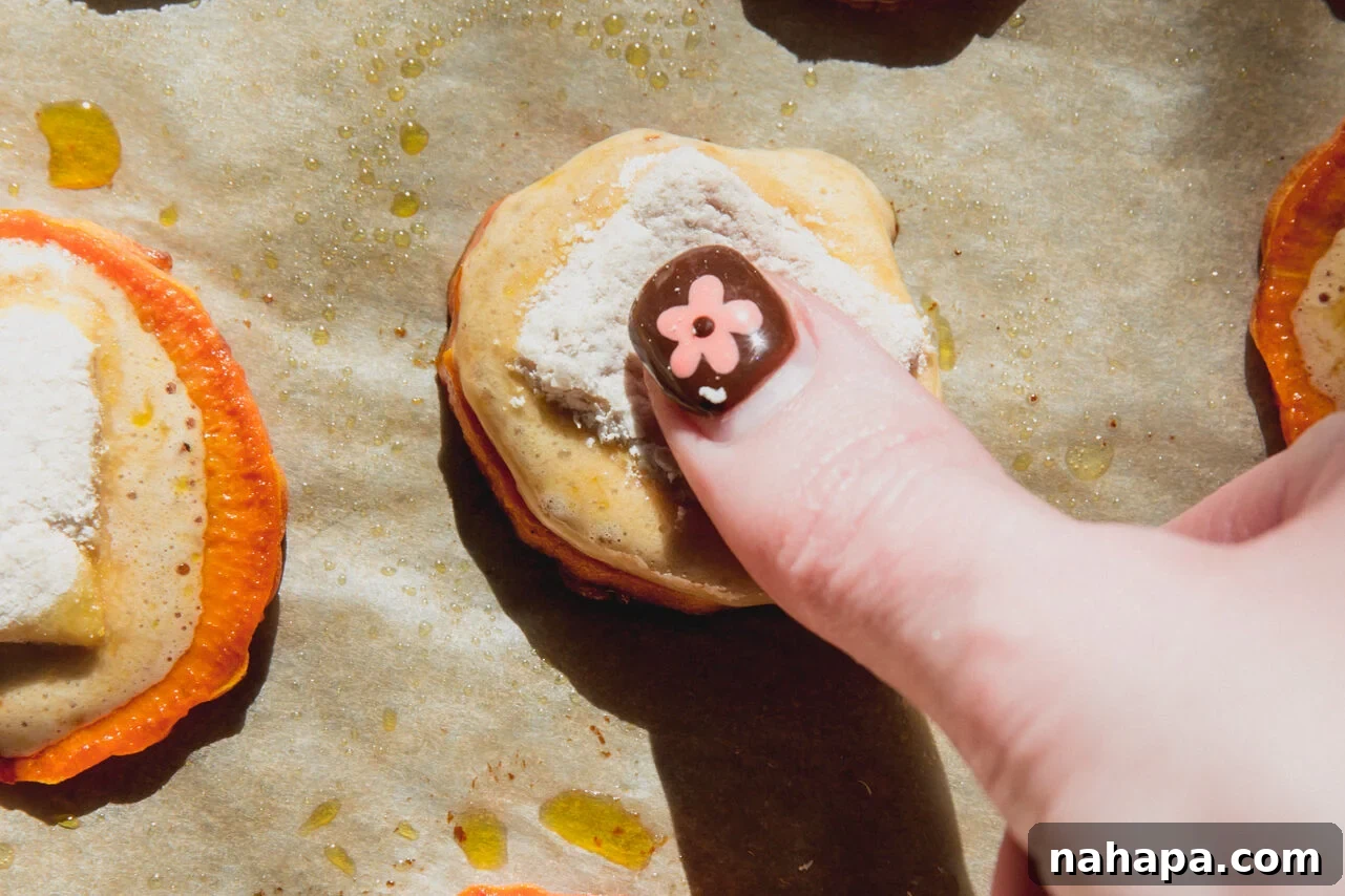 A thumb gently pressing a marshmallow into a warm sweet potato bite on a baking sheet.