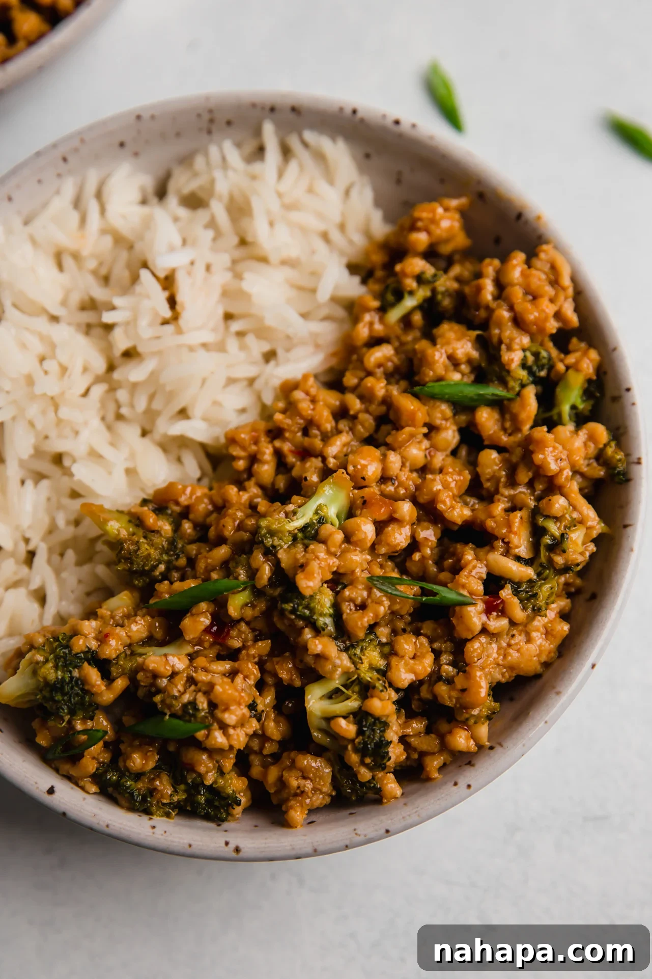 A vibrant bowl of firecracker ground chicken with crisp green broccoli florets, served alongside fluffy white rice, garnished with fresh chopped green onions. The setting suggests a comforting, homemade meal.