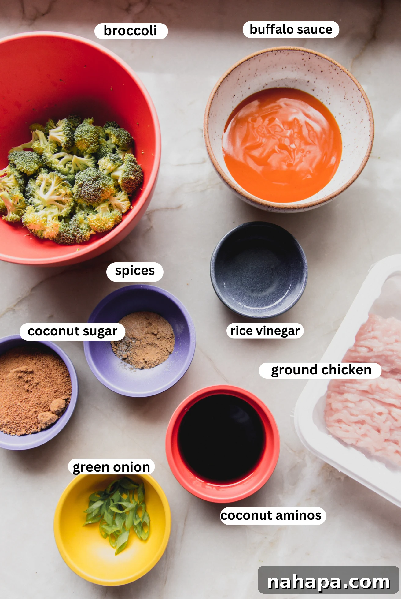 Individual ingredients for Firecracker Ground Chicken Bowls laid out in small white bowls, including buffalo sauce, coconut aminos, coconut sugar, a mix of spices, fresh broccoli florets, and raw ground chicken.