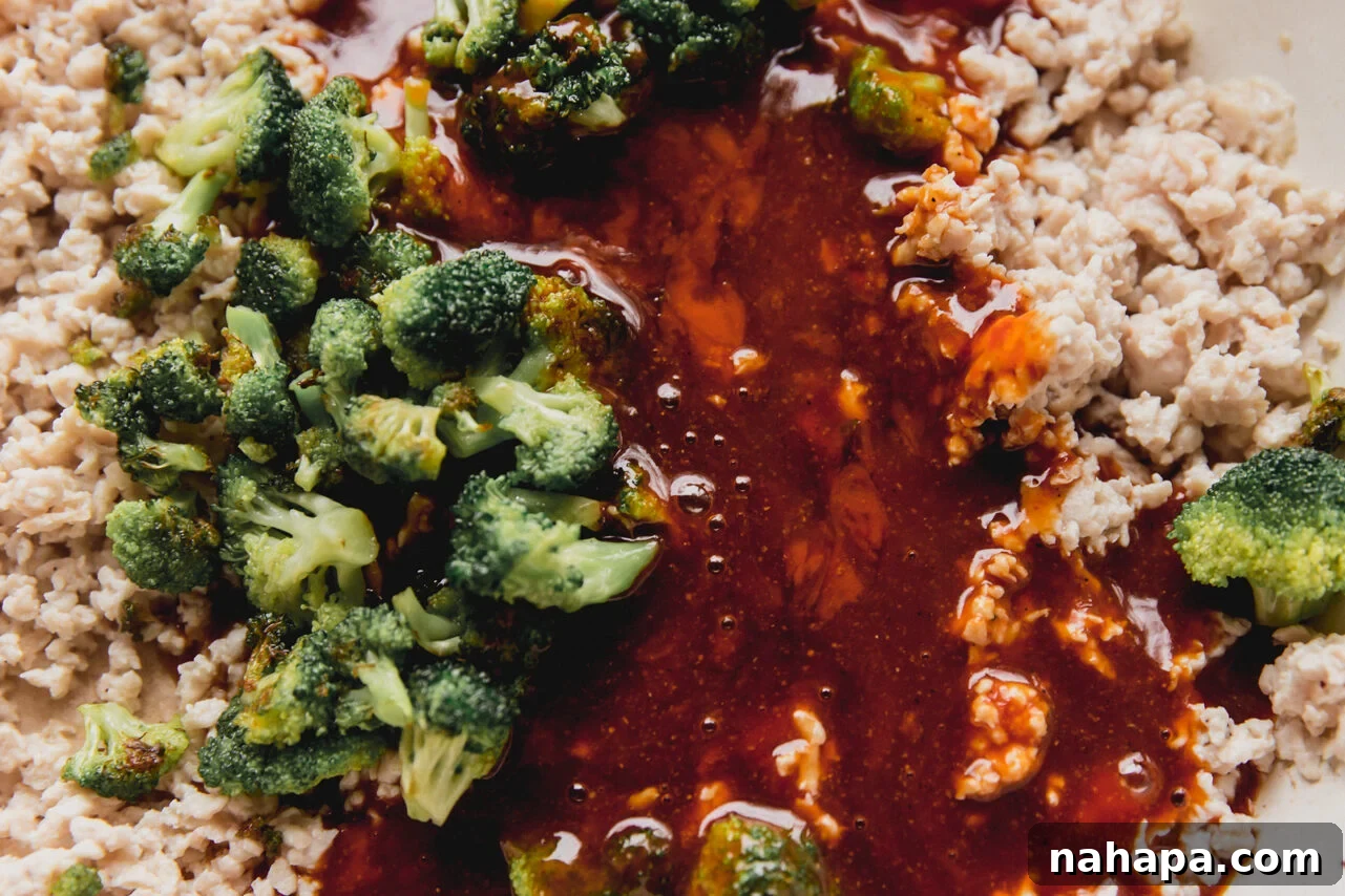 The cooked ground chicken and sautéed broccoli are combined in the pan, with the homemade firecracker sauce being poured generously over the mixture, ready to be stirred.