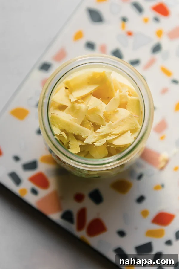 A clear glass jar filled with thinly sliced, peeled ginger, ready for the pickling brine.