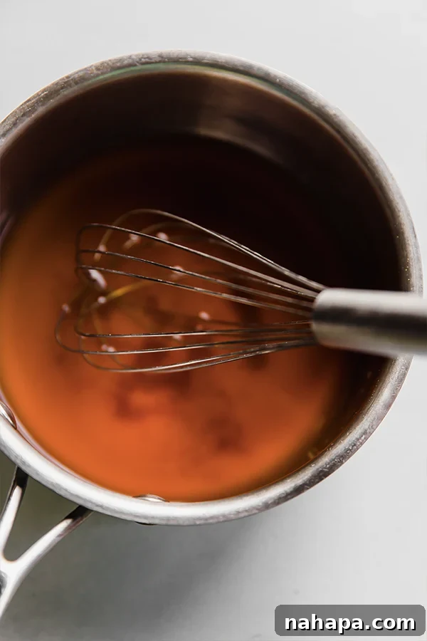A whisk stirring ingredients for pickled ginger in a saucepan on a stove, dissolving coconut sugar in the liquid.