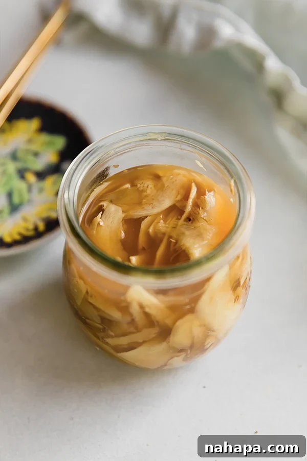 A clear glass jar filled with homemade pickled ginger, with a wooden spoon resting beside it, set against a rustic background.