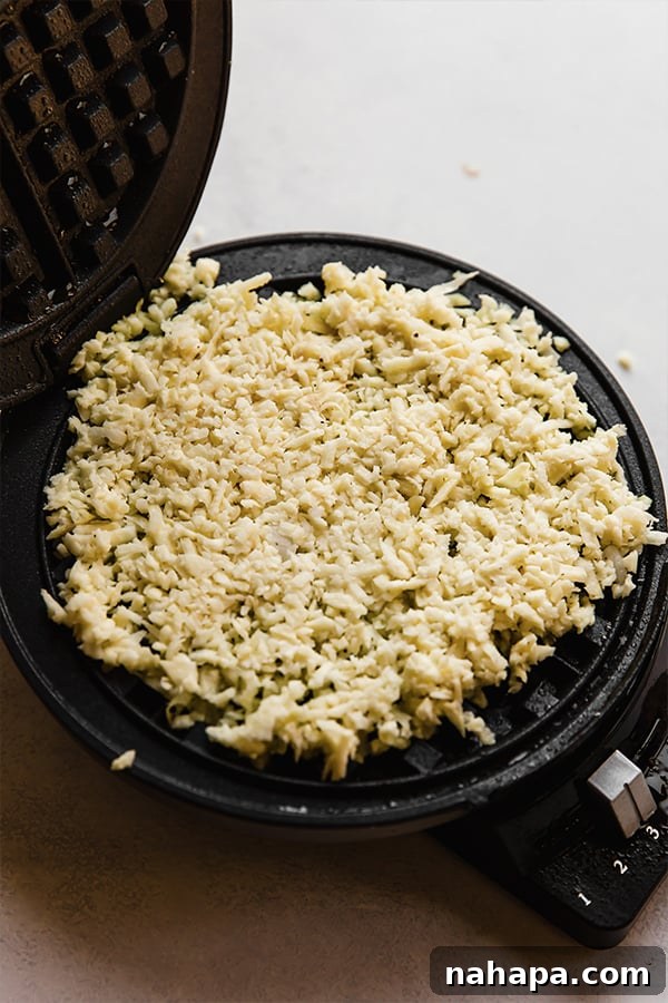 Shredded sweet potato mixture being pressed onto a hot, greased waffle griddle, starting to cook.