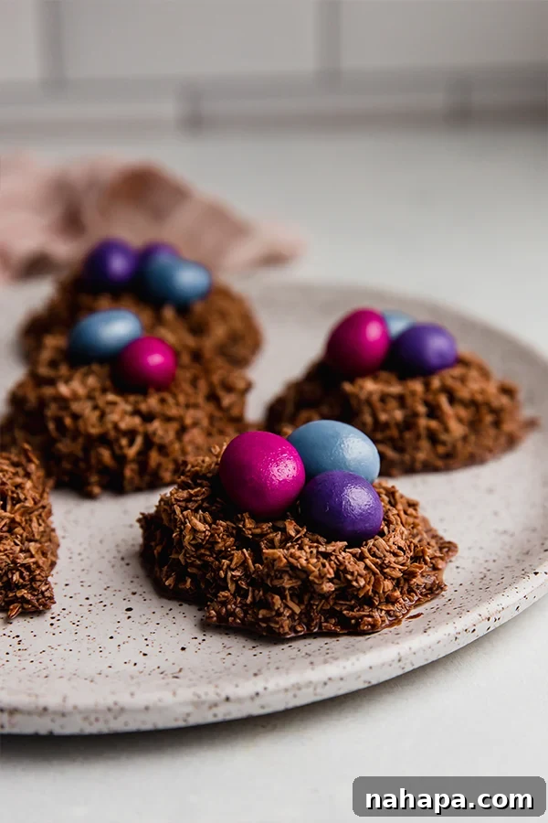 Sweet Coconut Chocolate Easter Nests 5 A close-up shot of multiple finished Chocolate Coconut Easter Egg Nests, each topped with colorful candy gems, arranged on a plate.