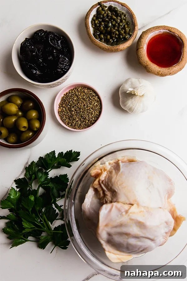 All the fresh ingredients for Chicken Marbella laid out on a table, including chicken, olives, capers, prunes, herbs, and oils.