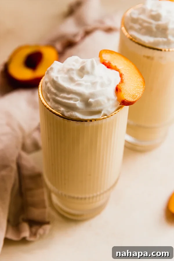 A vibrant, dairy-free creamy peach milkshake in a tall glass, garnished with fresh peach slices and a dollop of coconut whipped cream. It's an inviting and healthy summer treat.