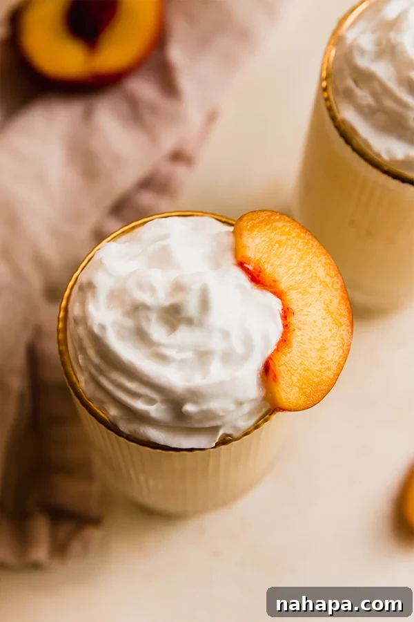 A tall glass of dairy-free creamy peach milkshake topped with coconut whipped cream and a few fresh peach slices, showcasing its inviting texture and presentation.