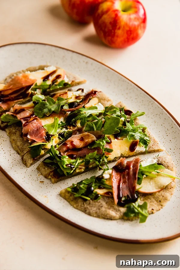A fully assembled and baked apple balsamic flatbread, garnished with fresh arugula, prosciutto, and balsamic glaze, presented on a serving plate.