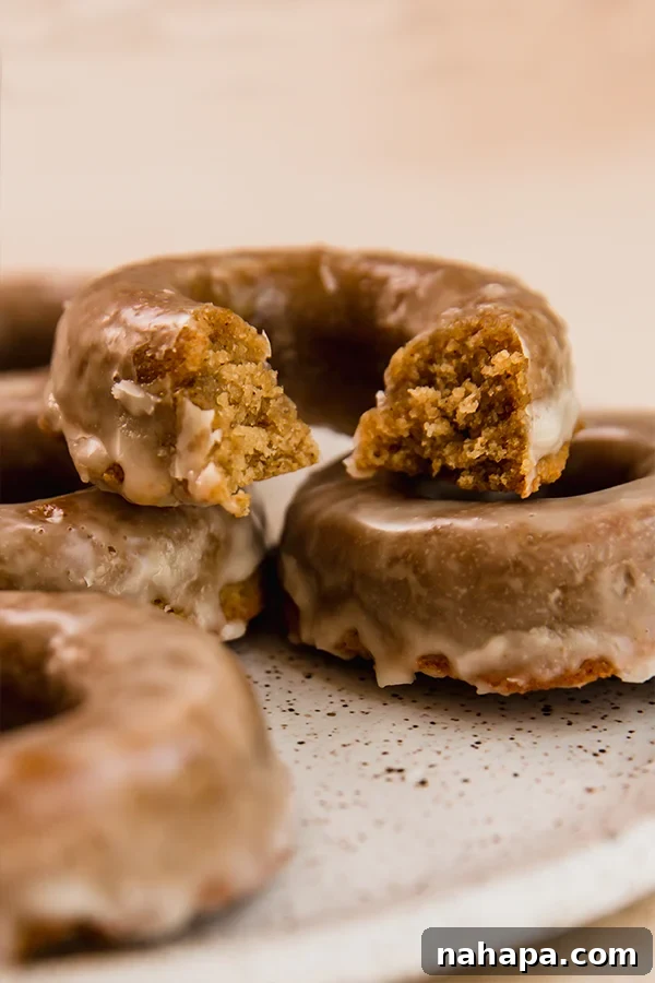 A stack of perfectly glazed apple cider donuts, with one having a bite taken out of it, showcasing its soft, spiced interior. These grain-free, paleo, and AIP-friendly treats are a fall favorite.