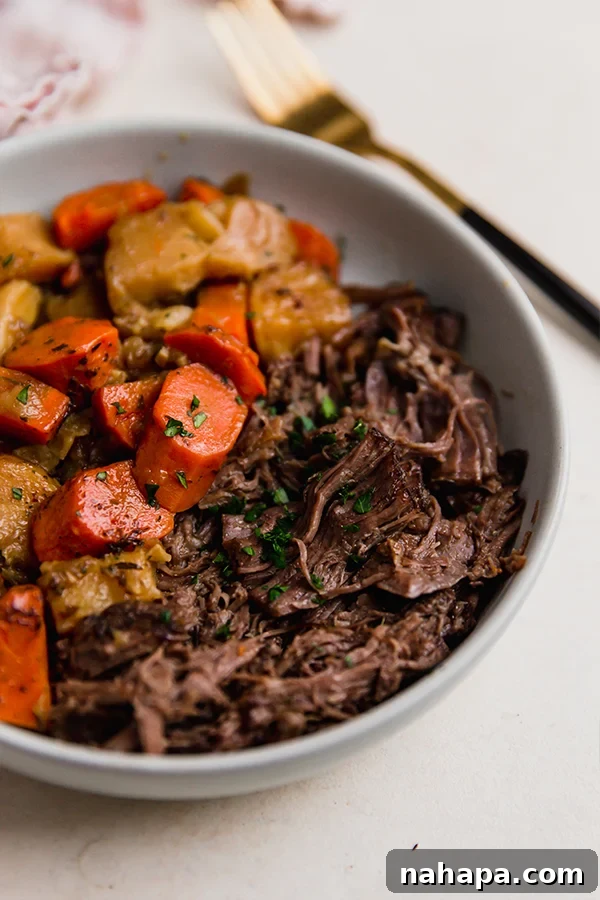 A close-up shot of a serving of shredded oven-baked chuck roast in a white bowl, garnished with fresh parsley and resting on a wooden surface.