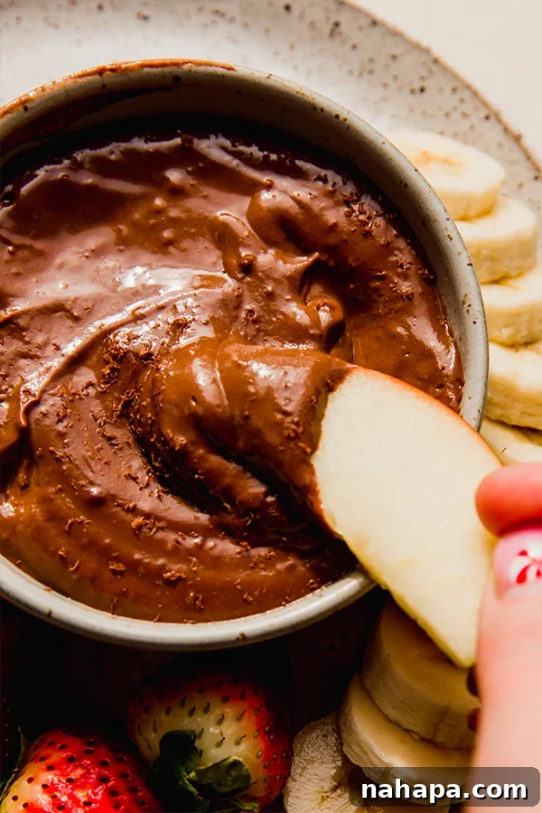 A crisp green apple wedge being dipped into a bowl of creamy, rich chocolate fruit dip, showcasing the luxurious texture of the dip.