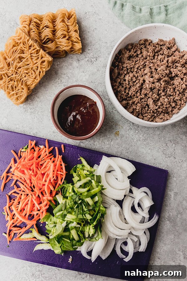 A collection of fresh ingredients laid out on a wooden surface, including dates, coconut aminos, rice vinegar, ground beef, gluten-free ramen, and various vegetables, ready for hoisin beef preparation.