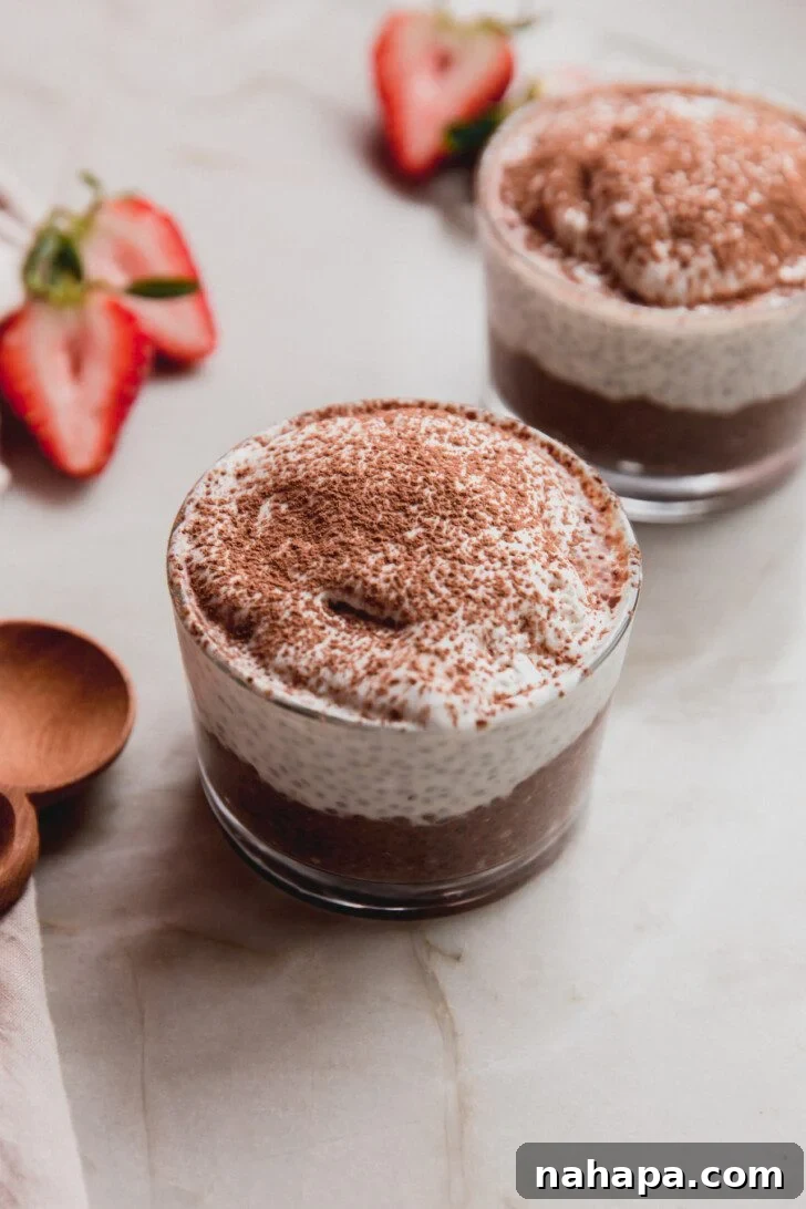 A small glass filled with beautifully layered tiramisu chia pudding, topped with fresh fruit and a dusting of cocoa, set against a softly blurred background.
