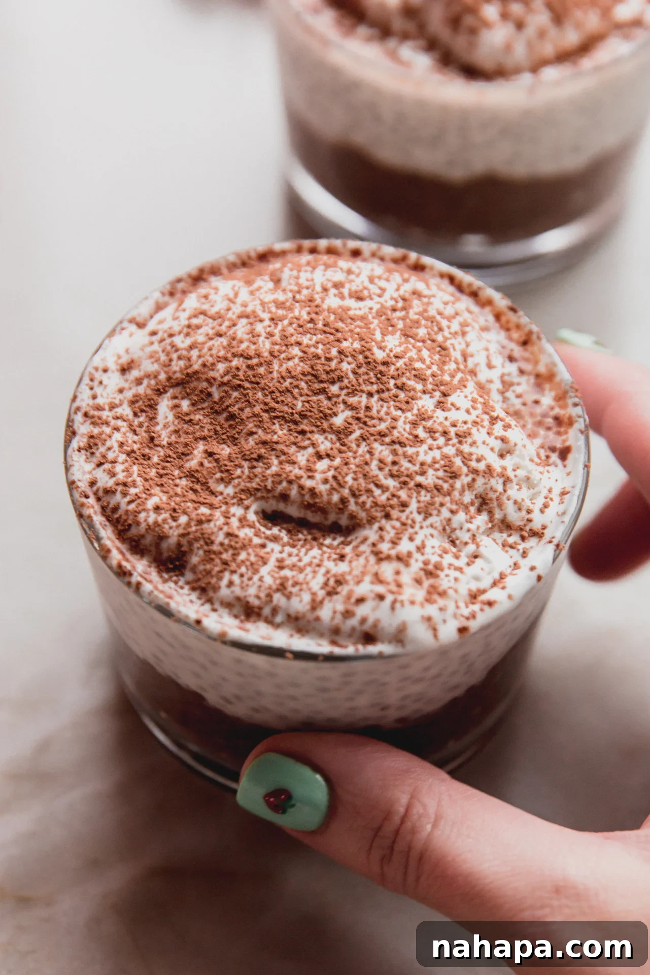 A hand holding a clear glass filled with layered tiramisu chia pudding, beautifully garnished with whipped cream and a dusting of cocoa powder.
