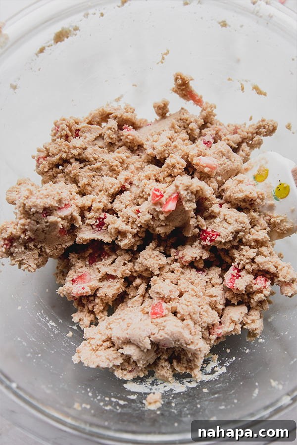 A close-up shot of the unbaked strawberry mini donut dough in a clear glass bowl, showcasing its thick, moist texture and the vibrant red flecks of diced strawberries.