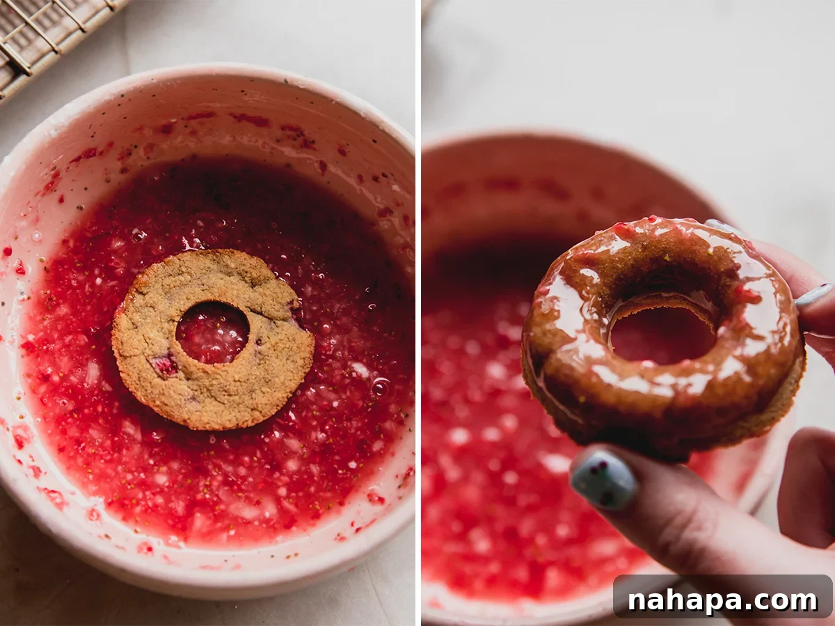 A close-up of a hand dipping a fluffy strawberry mini donut into a vibrant pink strawberry glaze, showcasing the smooth, luscious coating.